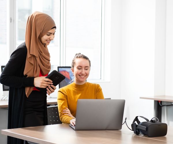 Students in a Media Production and Design classroom working on a laptop; a VR headset sits next to them on a desk.