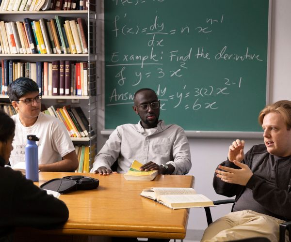 Students and a professor having a discussion at a table; a chalkboard with mathematical equations is displayed in the background.