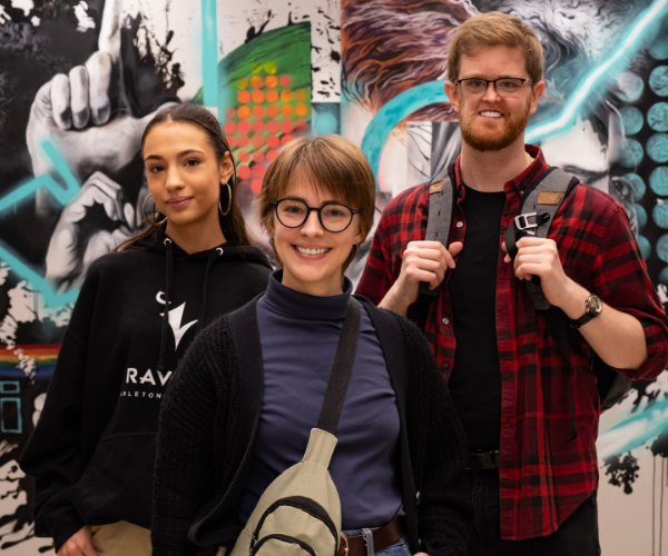 Three students standing in front of a wall. The wall has a collage of paintings on it.It is located in Dunton Tower in the Social Work section of Carleton University.