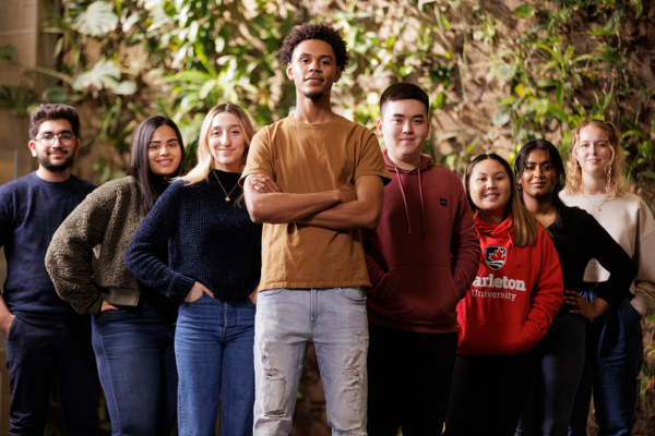 Carleton students standing in front of the green wall, it is a so-called living wall covered with plants. There are 7 students and they are all looking at the camera.