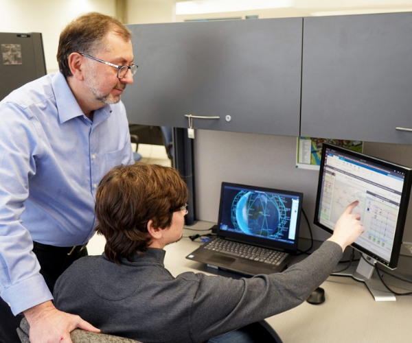 Student working with professor. The student is pointing at a desk top and the professor is standing behind him looking toward the desktop. 