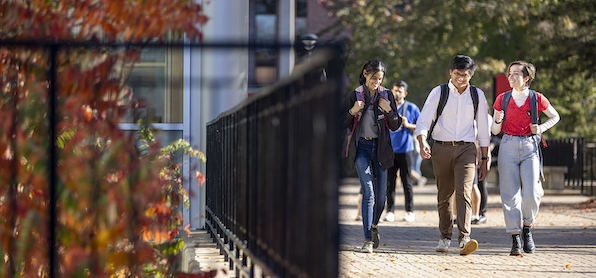 three students walking on a pathway outdoors on campus, by a fence, trees all around