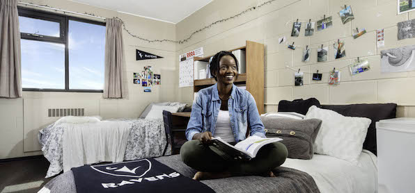 Girl sitting crossed legged on bed in residence room, pictures on the wall, book on her lap