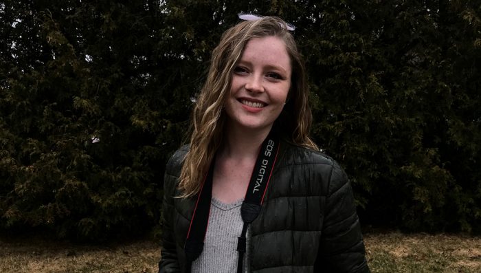 Student Sarah MacFarlane standing in front of trees with camera.