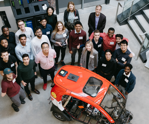 photo of a group at the capstone engineering fair. The group of students are standing in a half-circle around a smart car that they created. 